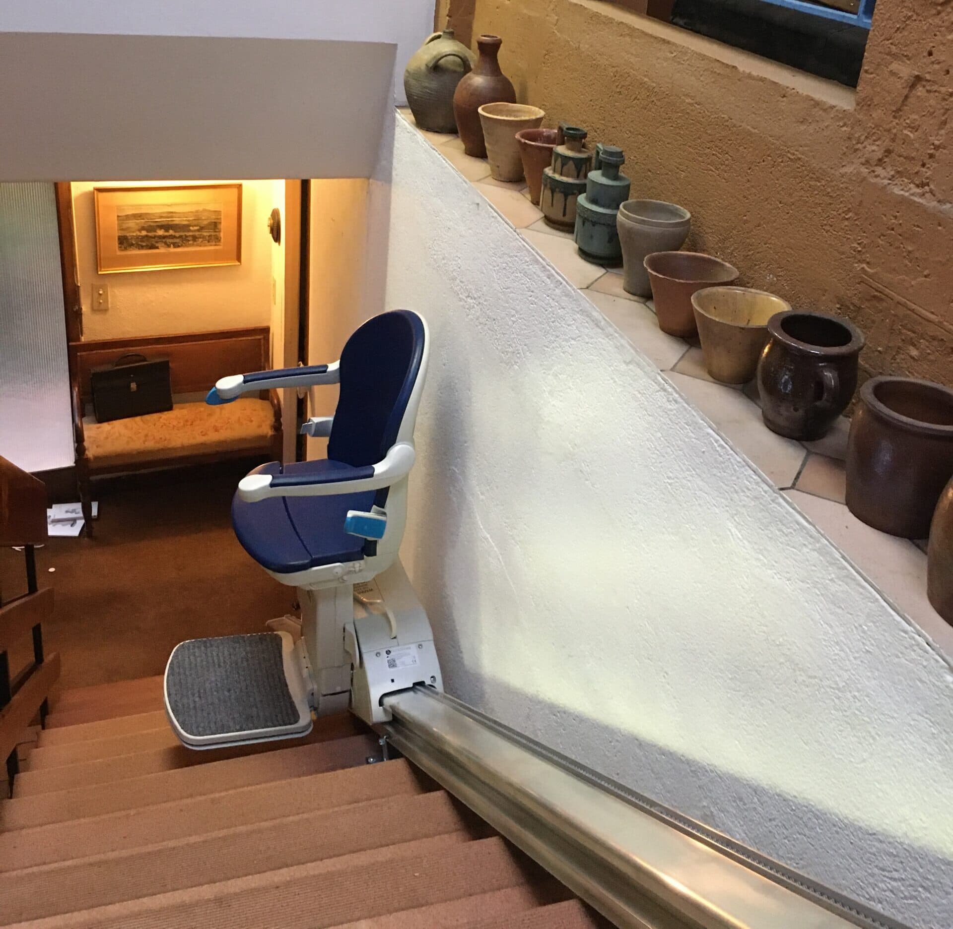 Monte-escalier à siège bleu replié en haut d'un escalier, dans une maison avec des poteries